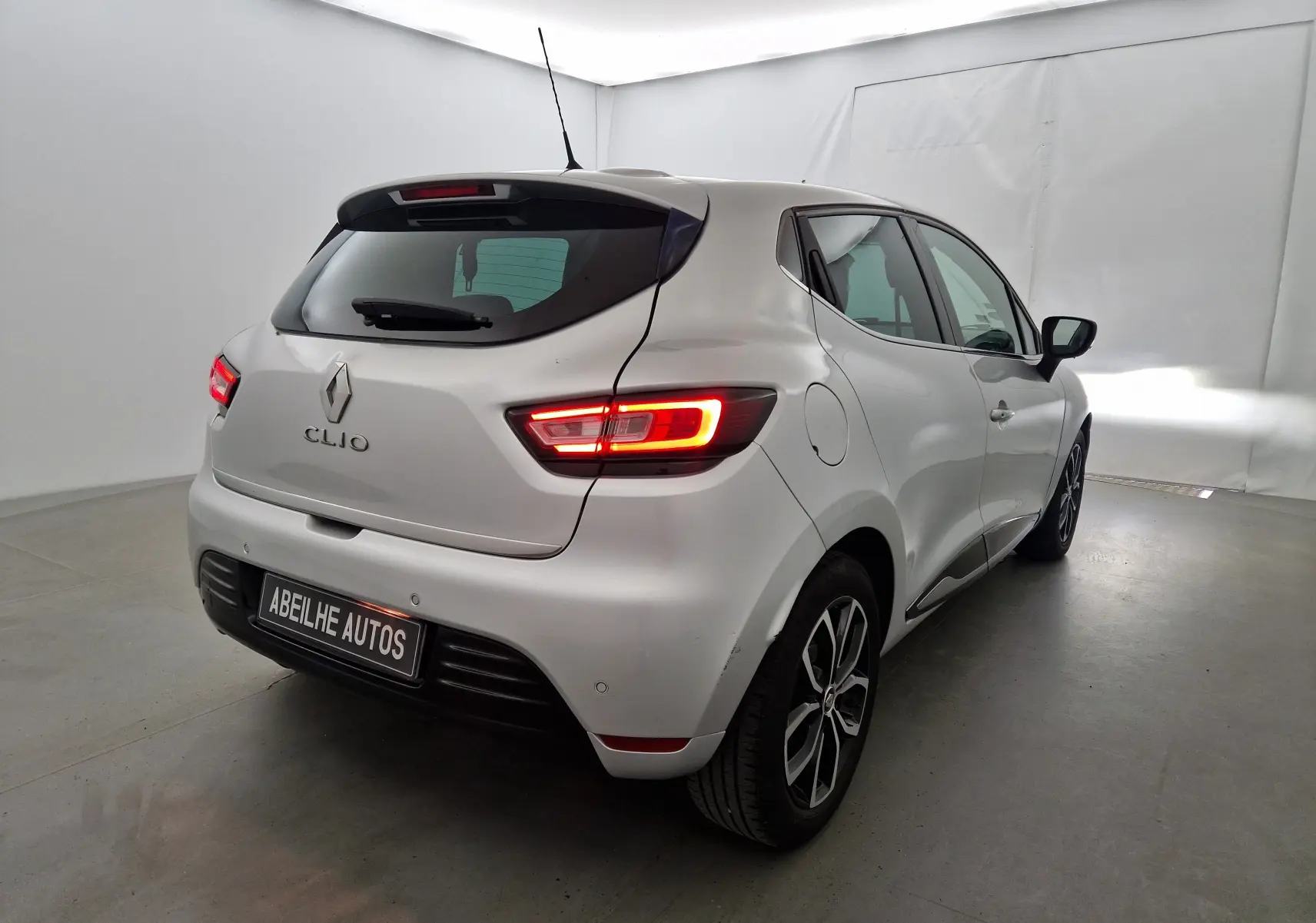 Vue 3/4 arrière droite d'une Renault Clio IV gris clair métal avec feux arrière allumés dans un studio blanc.