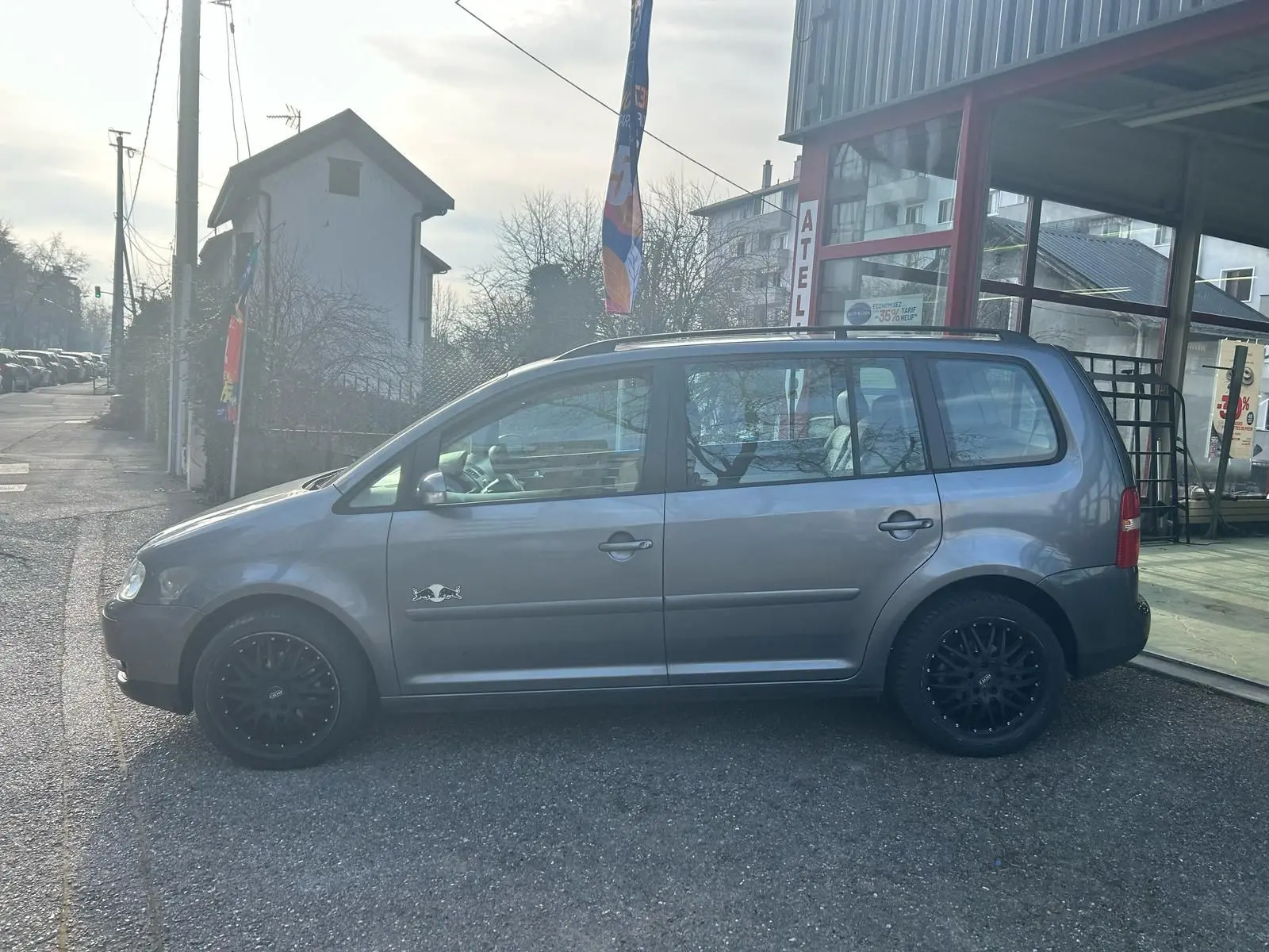 Profil côté gauche d'un Volkswagen Touran gris 2004 avec jantes noires et logo Red Bull sur la porte avant.