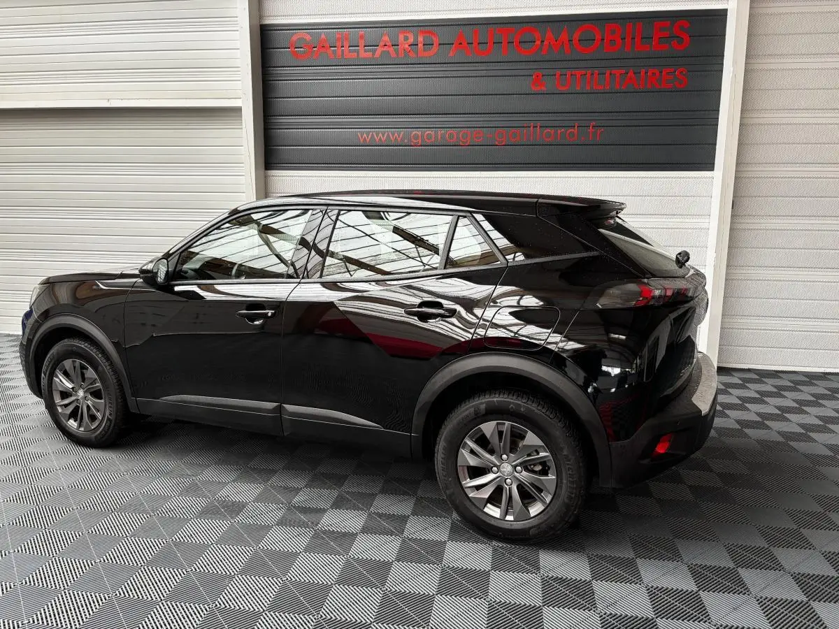 Peugeot 2008 noir vue de profil côté gauche dans un garage avec sol à damier et panneau publicitaire en arrière-plan.