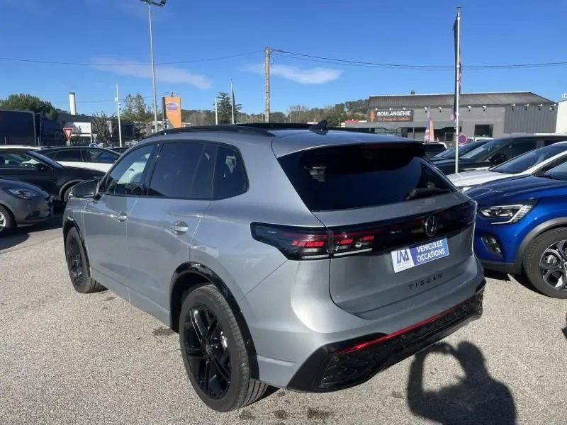 Vue 3/4 arrière droite d’un Volkswagen Tiguan 1.5 eHybrid gris métal avec finition R-line et jantes noires sur parking.
