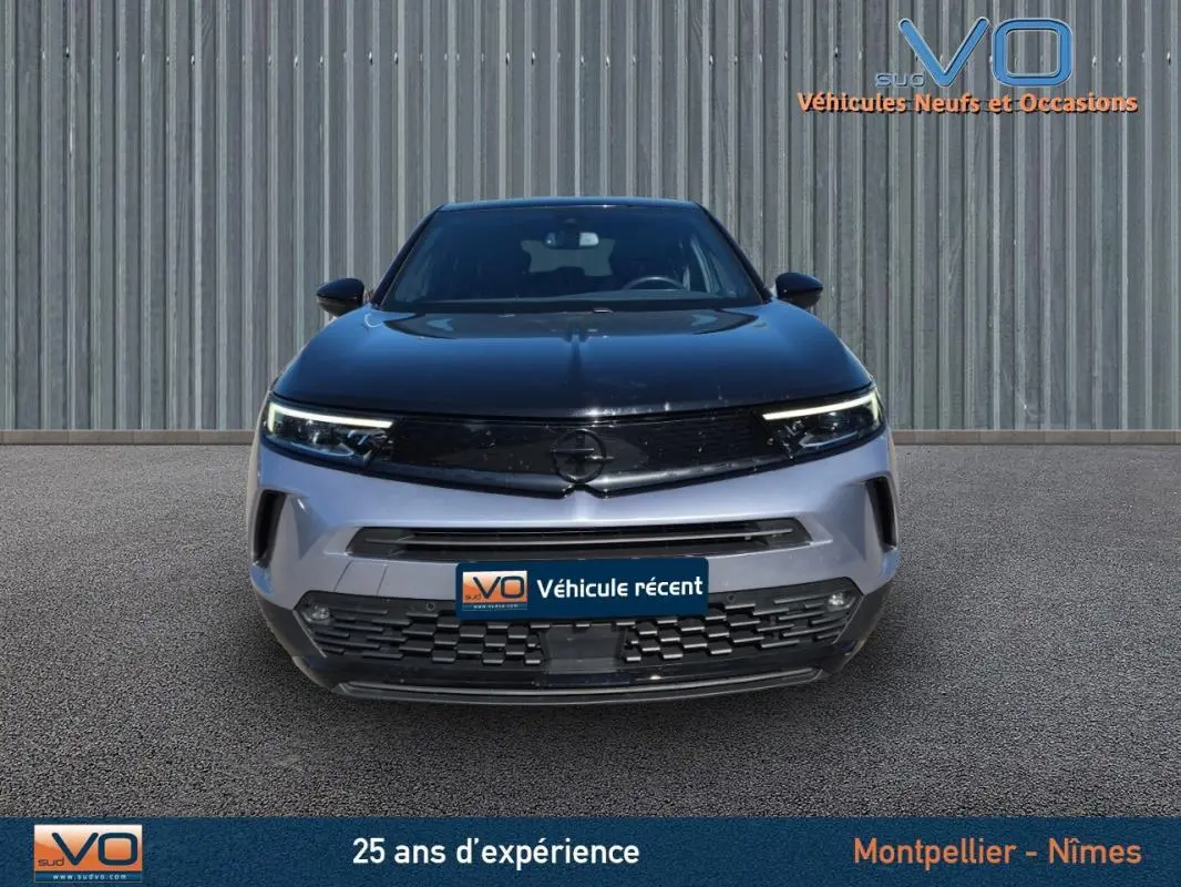 Vue frontale d'un Opel Mokka gris clair 2022 avec calandre noire et phares LED allumés sur fond industriel.