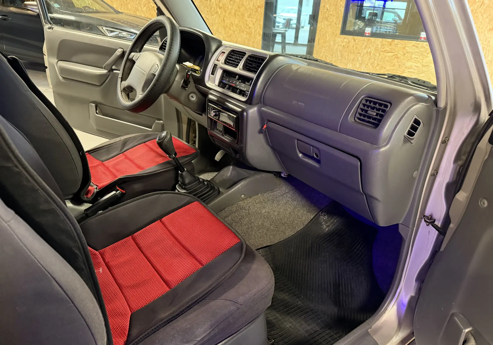 Vue intérieure côté passager du Suzuki Jimny Cabriolet gris de 2005, avec sièges avant noirs et rouges et tableau de bord simple.