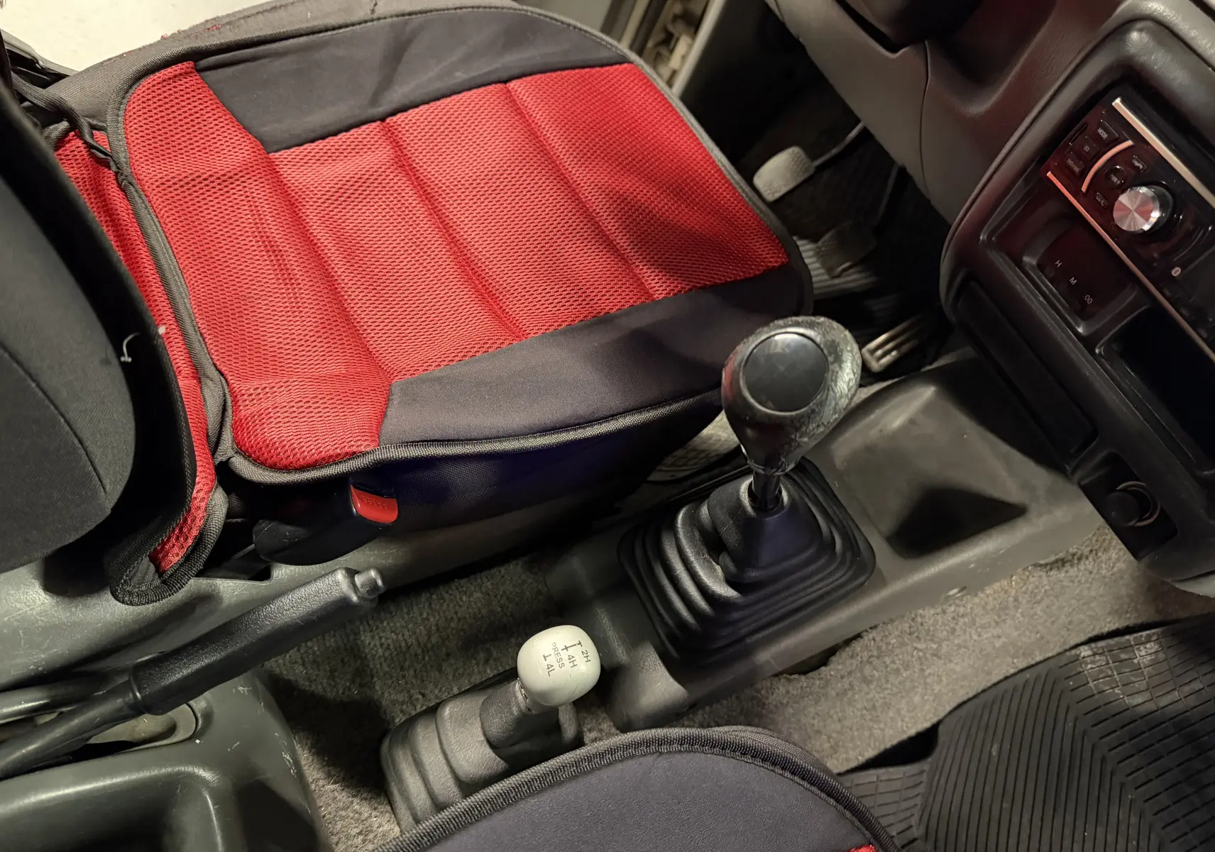 Vue intérieure centrée sur la console centrale d’un Suzuki Jimny Cabriolet gris, montrant levier de vitesse manuel et siège avec coussin rouge.