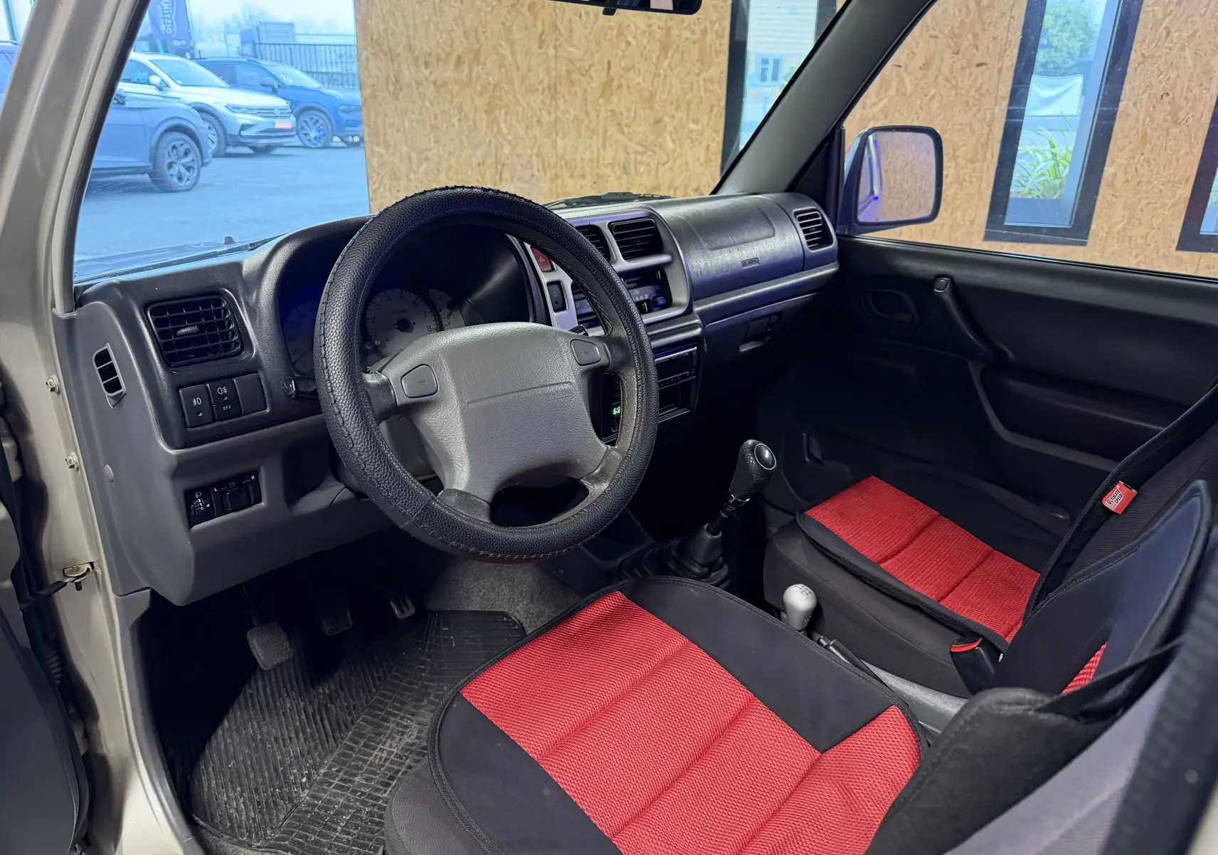 Vue intérieure côté conducteur du Suzuki Jimny Cabriolet gris 2005, volant, tableau de bord et sièges rouges visibles.