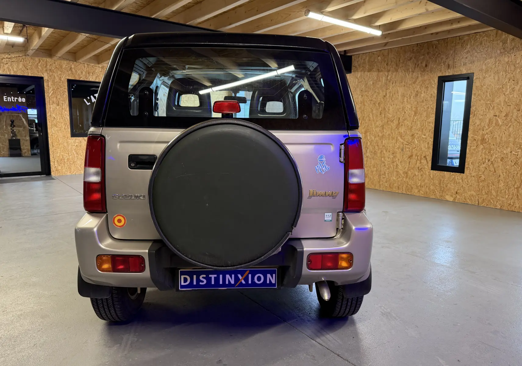 Vue arrière d’un Suzuki Jimny Cabriolet gris de 2005 avec roue de secours protégée et hard-top en intérieur.