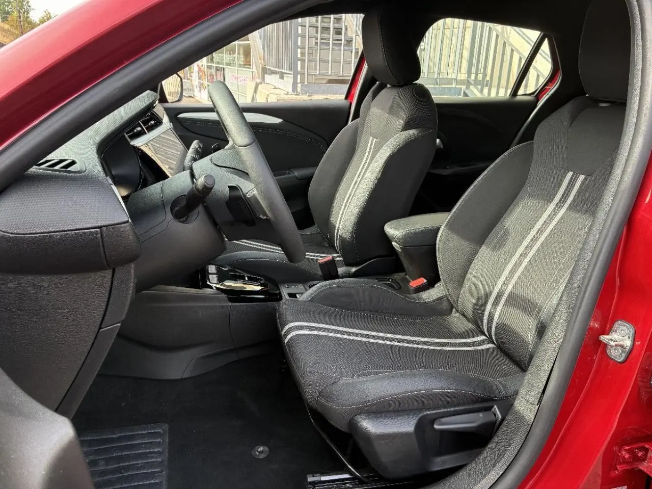 Vue intérieure côté conducteur de l’Opel Corsa 2025 rouge peperoncino, avec sièges noirs à bandes grises et tableau de bord moderne.