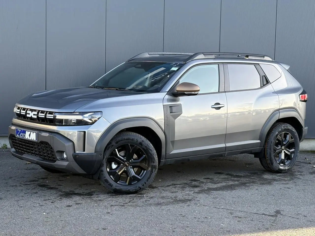 Dacia Duster 2026 gris schiste en vue 3/4 avant droit, avec jantes alliage noires et barres de toit longitudinales.