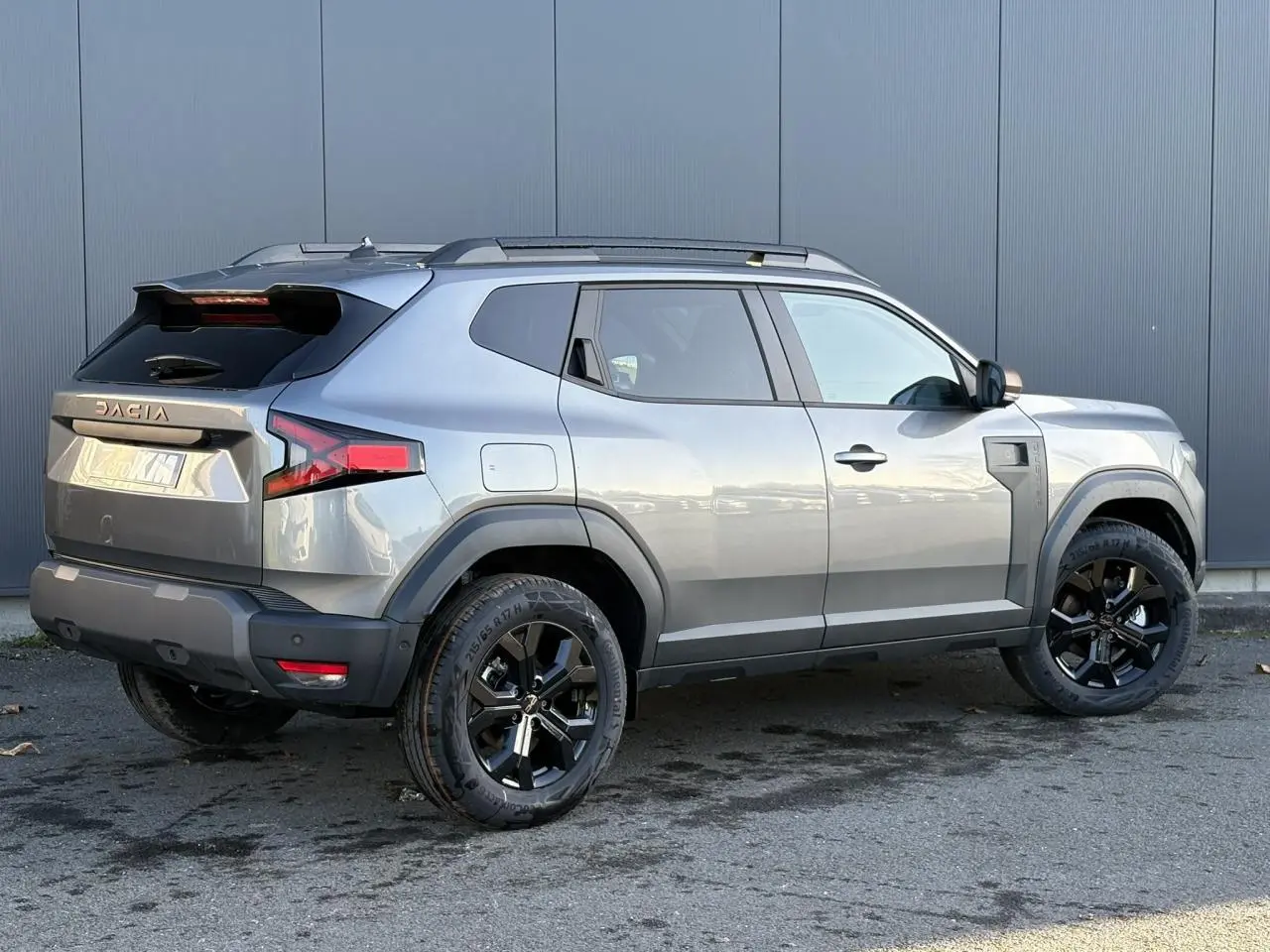 Vue 3/4 arrière droite du Dacia Duster 2026 gris schiste avec jantes alliage noires et barres de toit longitudinales.