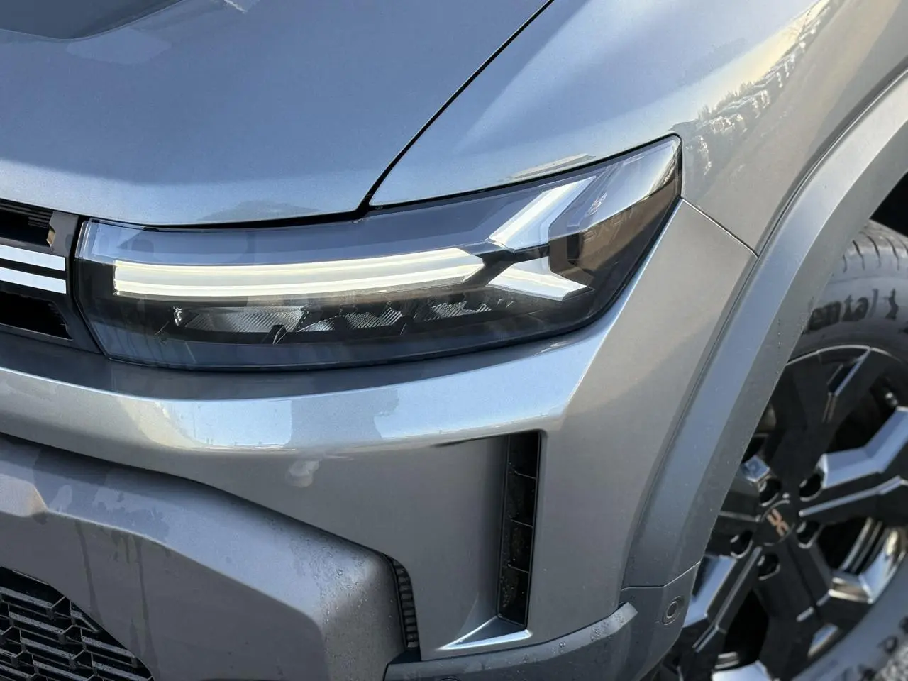 Gros plan sur le phare avant droit et la calandre du Dacia Duster gris schiste avec jante alliage noire visible.