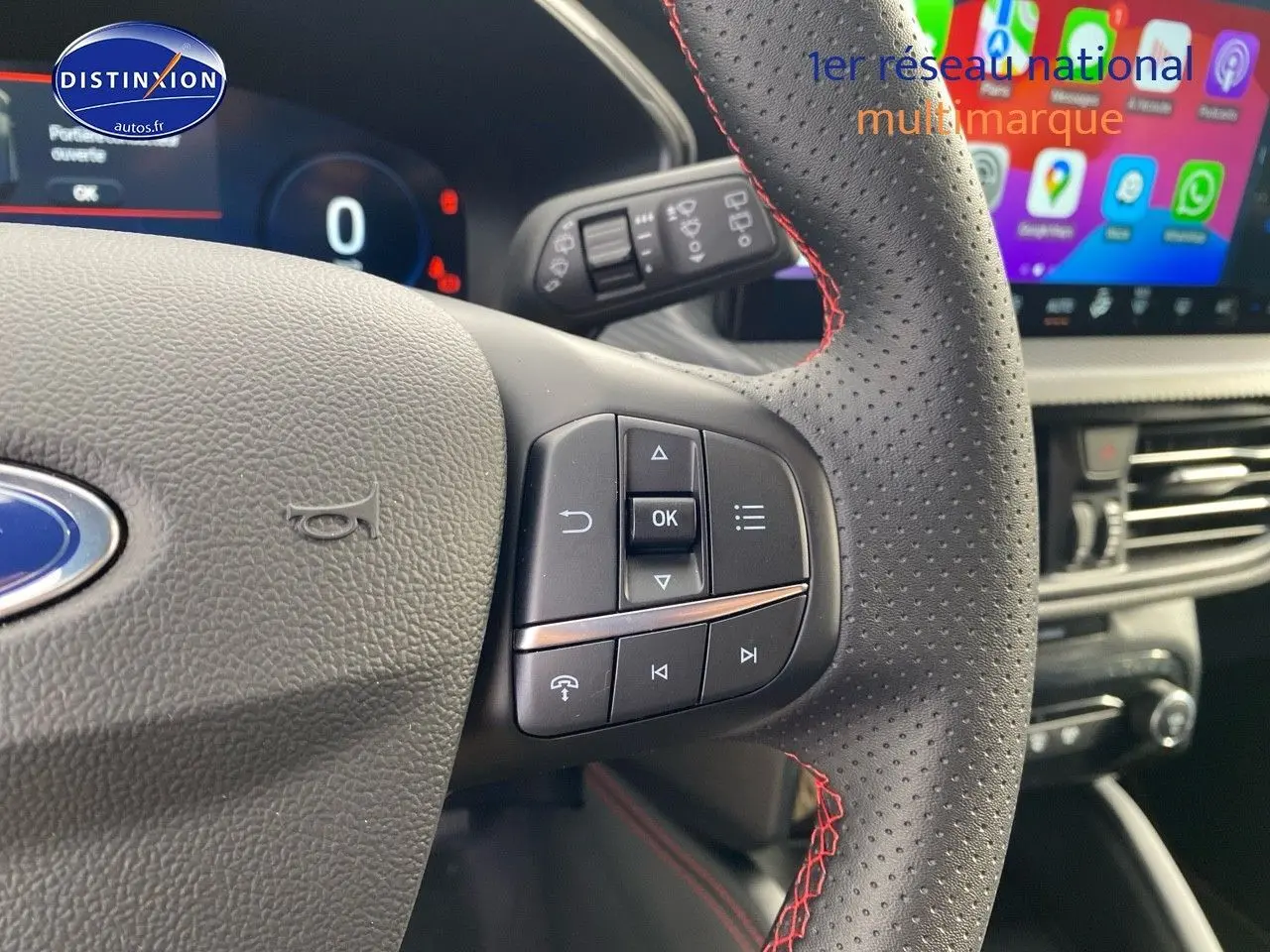 Gros plan sur le volant perforé avec surpiqûres rouges du Ford Kuga 2025, écran tactile et tableau de bord numériques visibles.