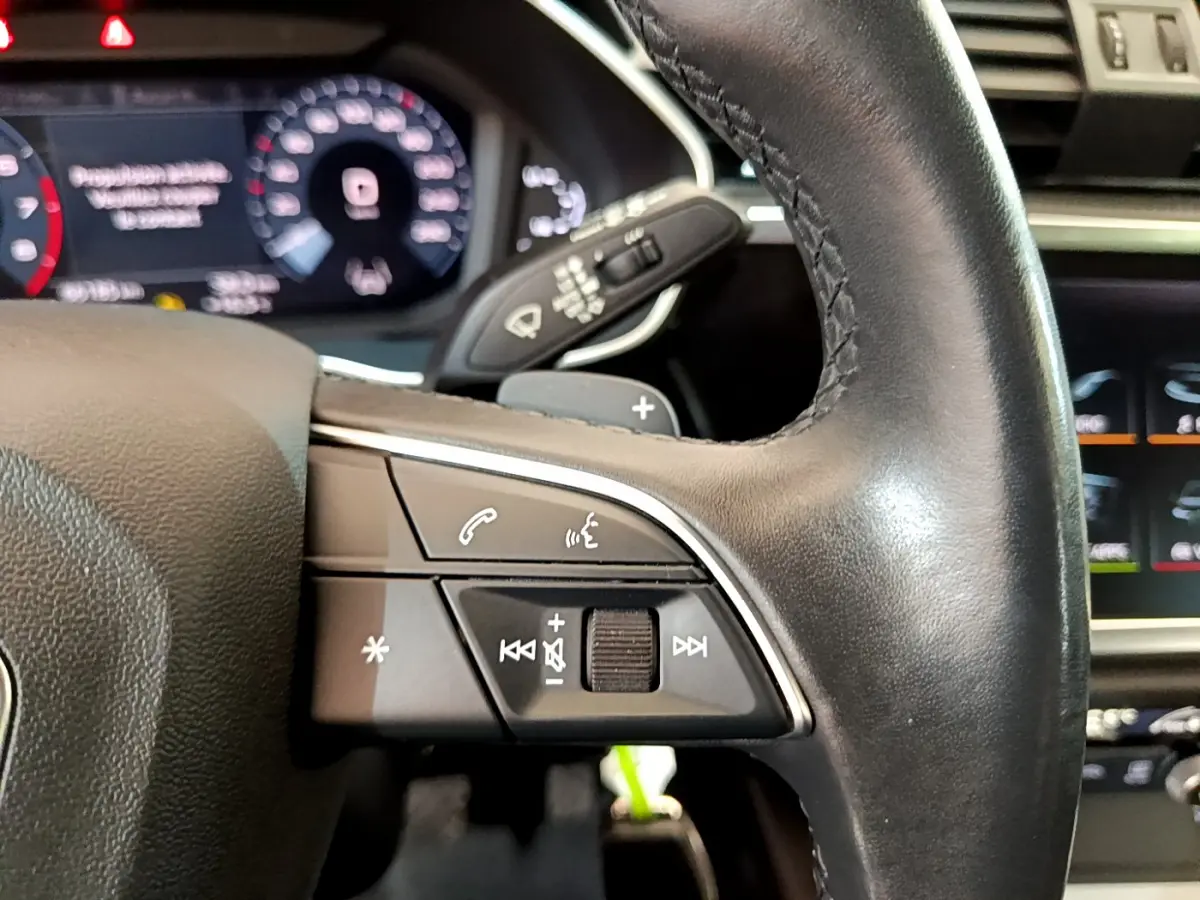 Gros plan sur le volant cuir noir de l'Audi Q3 Sportback 35 TFSI, avec commandes audio et affichage numérique en arrière-plan.