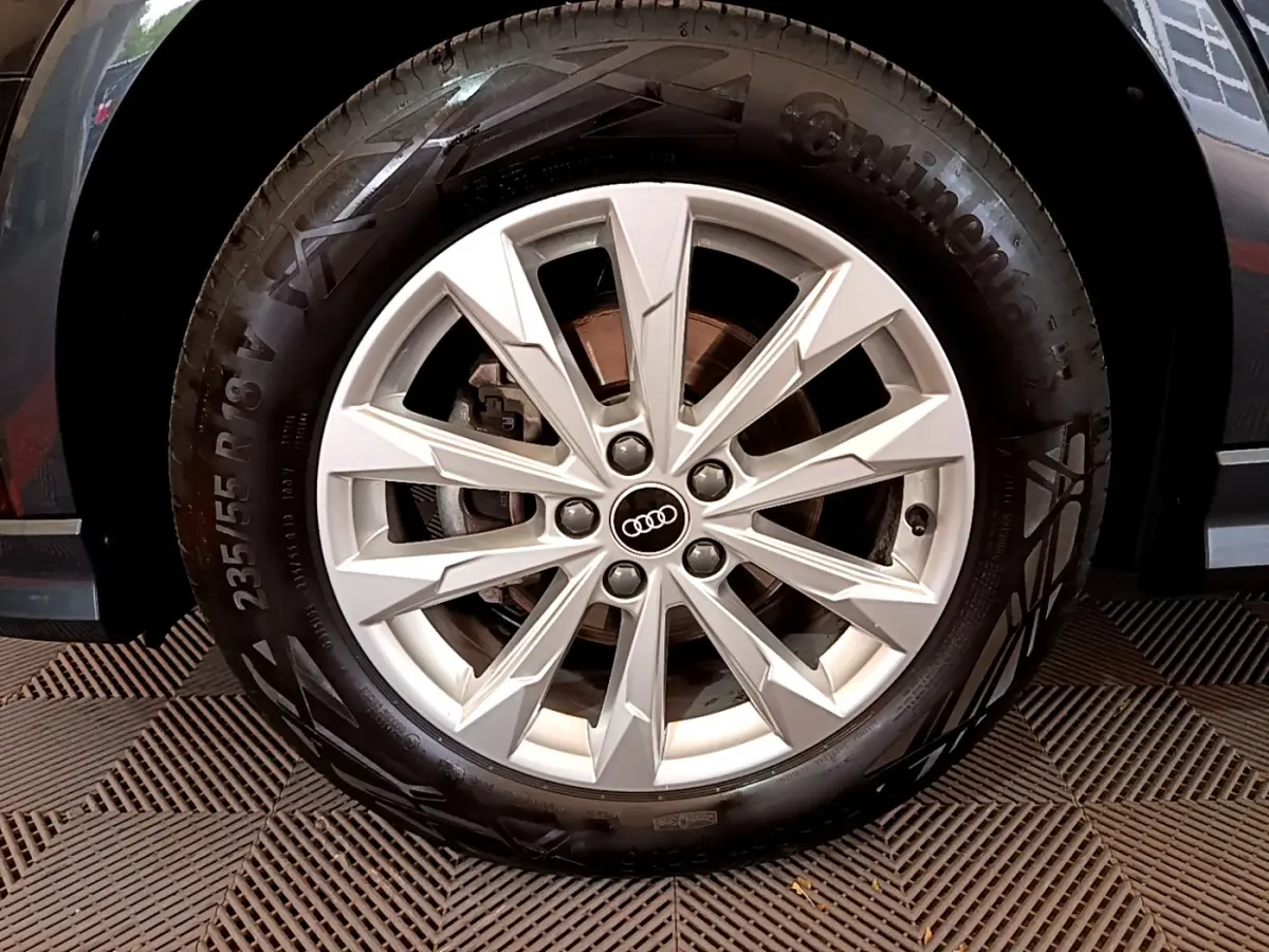 Gros plan sur la roue avant droite avec jante argentée et logo Audi, partie noire de la carrosserie visible en arrière-plan.