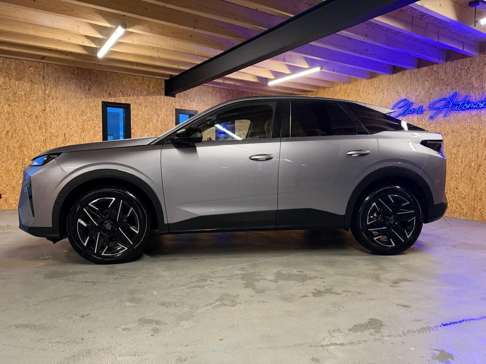 Vue de profil côté gauche du Peugeot 3008 gris 2025 avec jantes noires et toit noir dans un intérieur en bois.