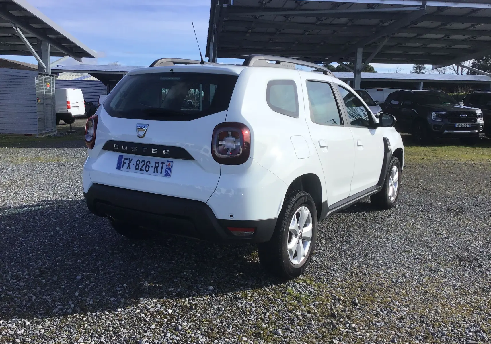 Vue 3/4 arrière droite d'un Dacia Duster blanc 2021 avec jantes alliage et barres de toit sous un abri.