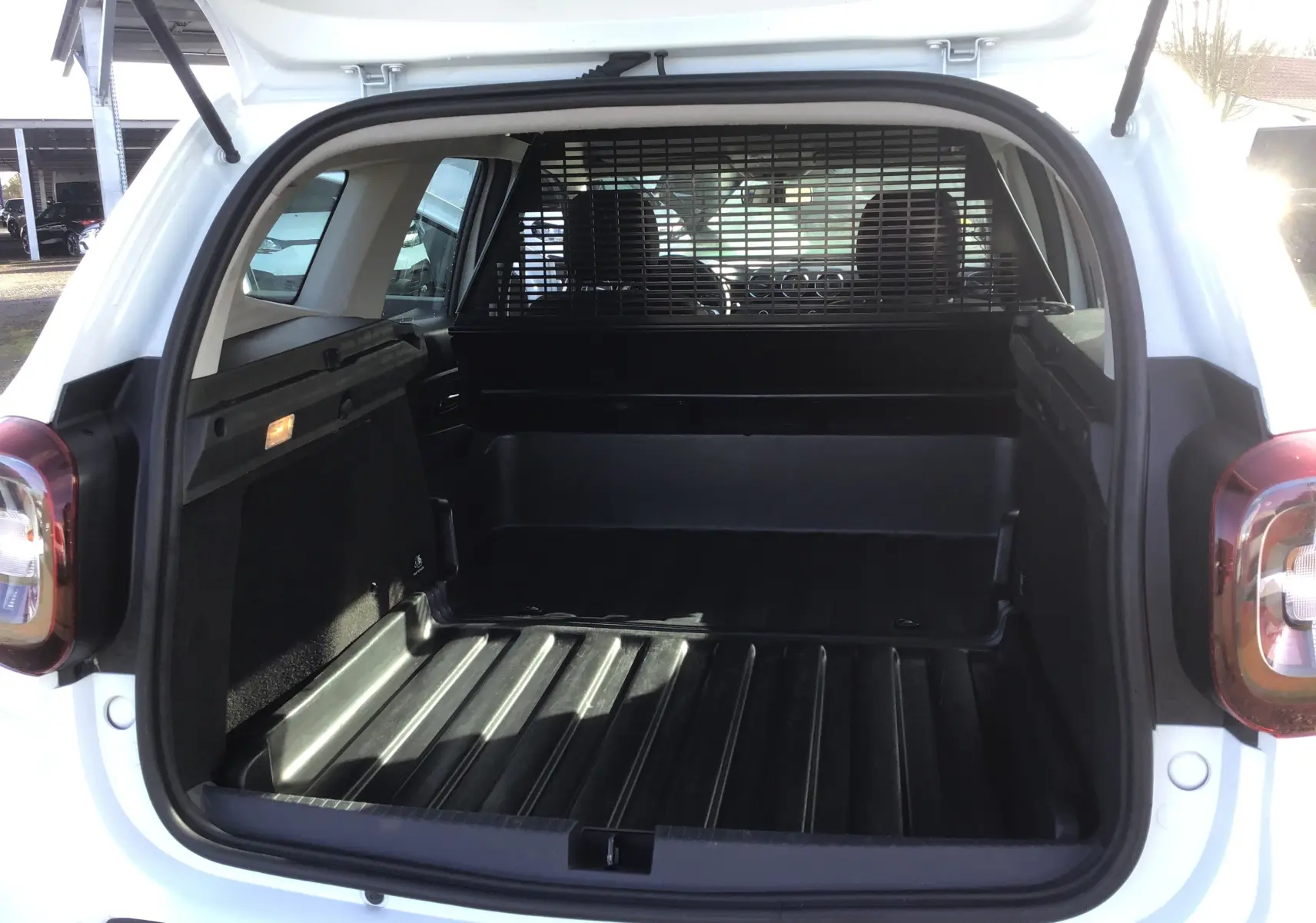 Vue arrière du coffre ouvert d'un Dacia Duster blanc 2021 avec grille de séparation et revêtement plastique noir.