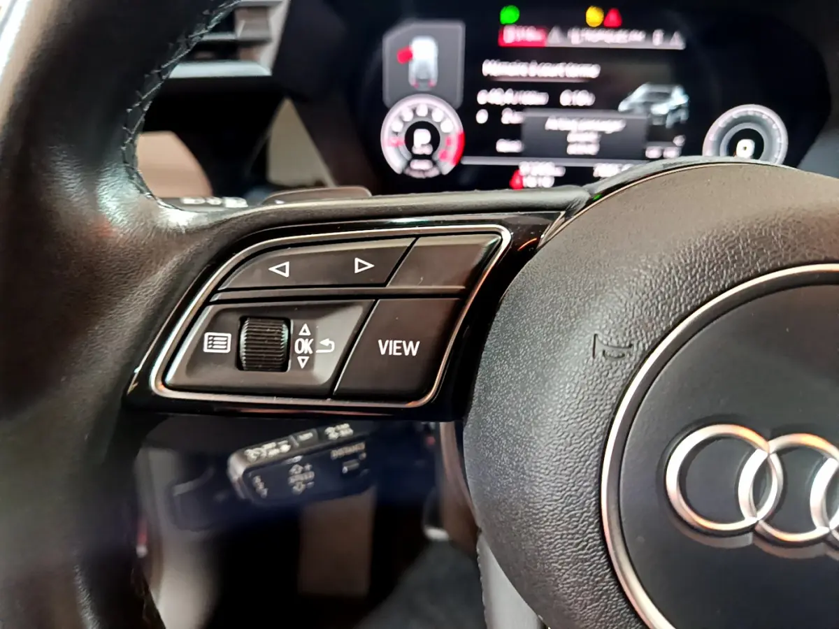 Gros plan sur le volant cuir noir de l'Audi A3 bleu 2023, avec commandes multifonctions et tableau de bord numérique flou.