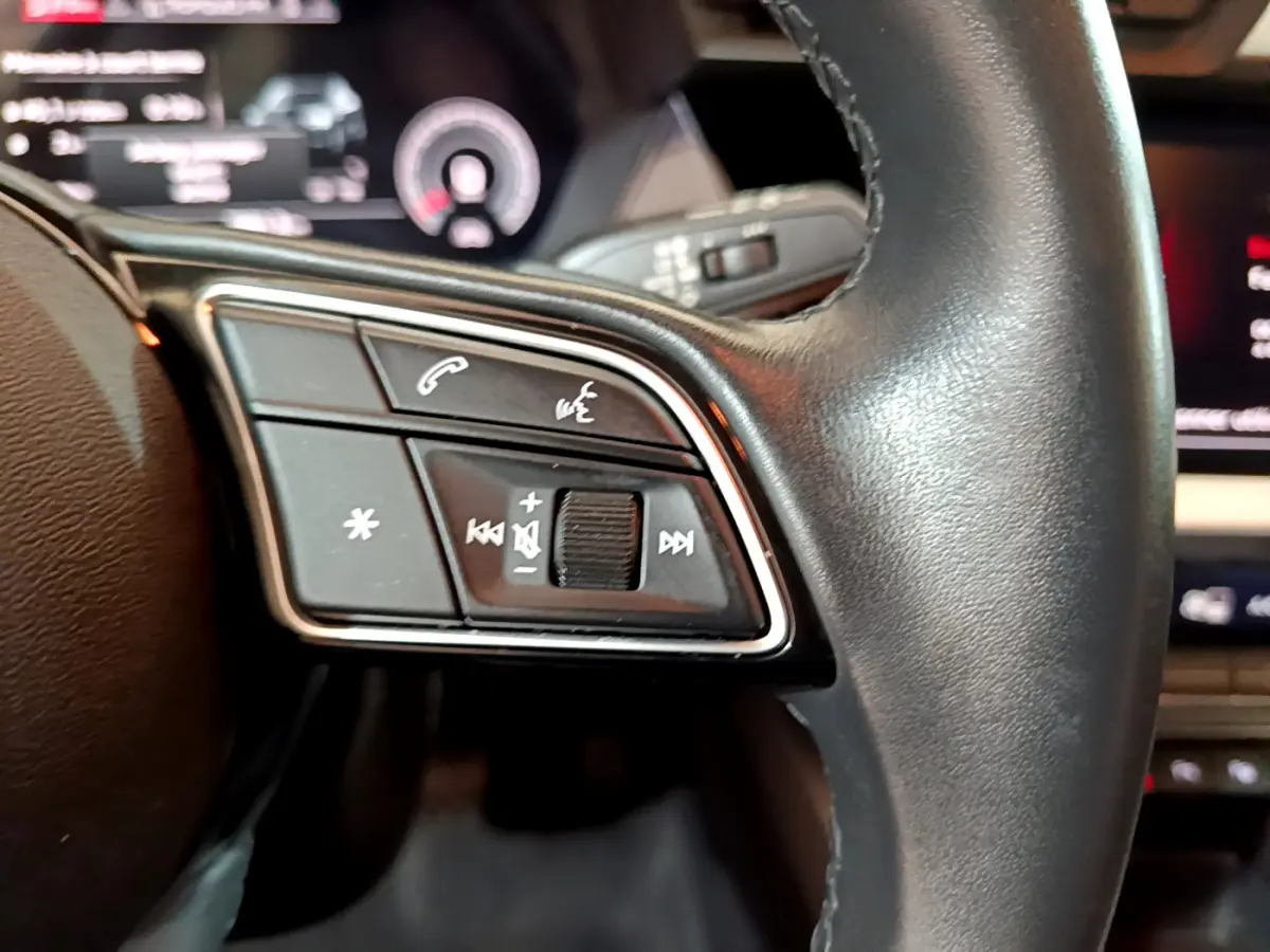 Gros plan sur les commandes gauche du volant cuir de l'Audi A3 35 TFSI bleu, avec affichage numérique en arrière-plan.