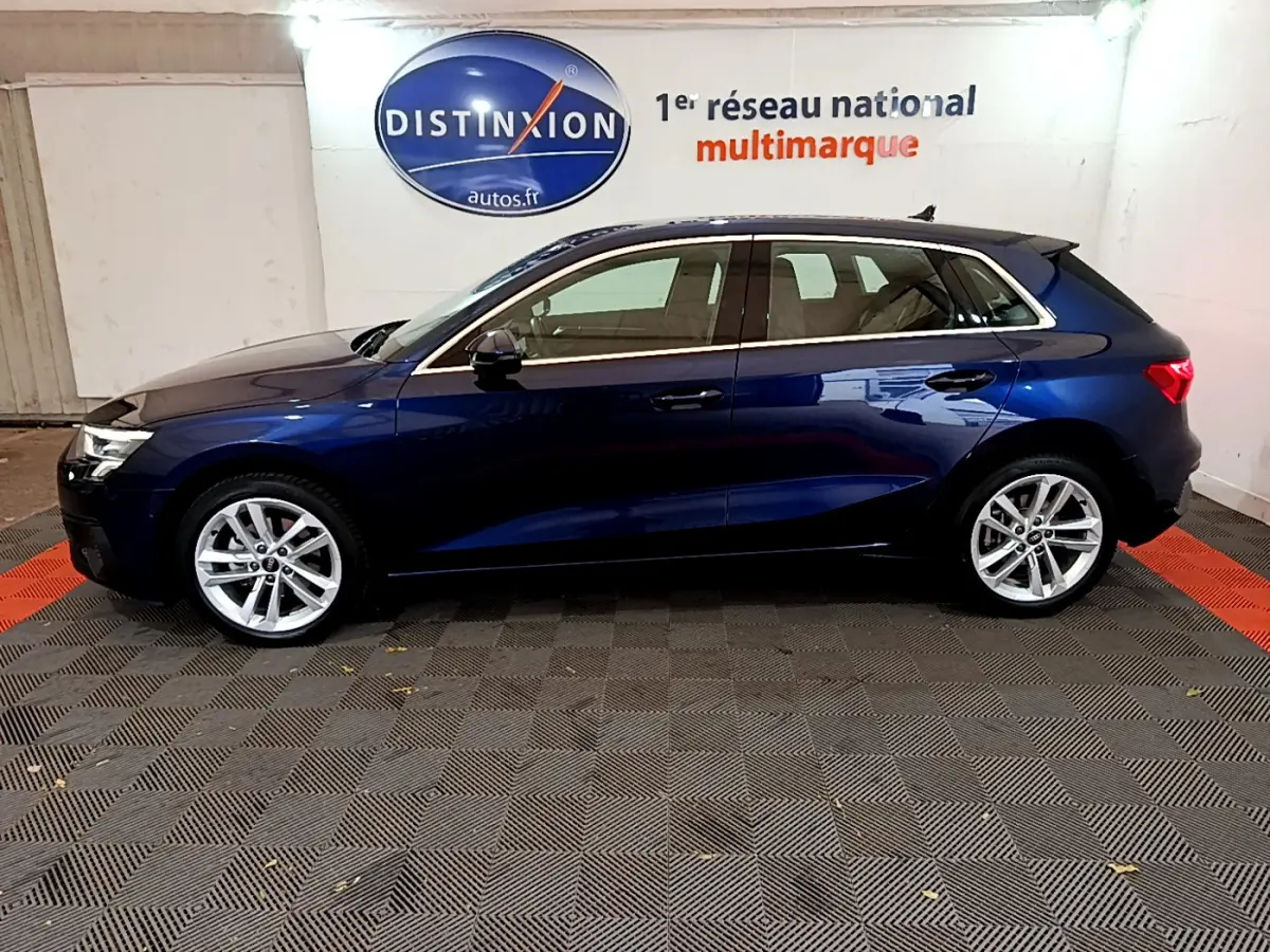 Profil côté gauche d'une Audi A3 35 TFSI hybride bleu foncé, stationnée en intérieur sur sol damier noir et rouge.