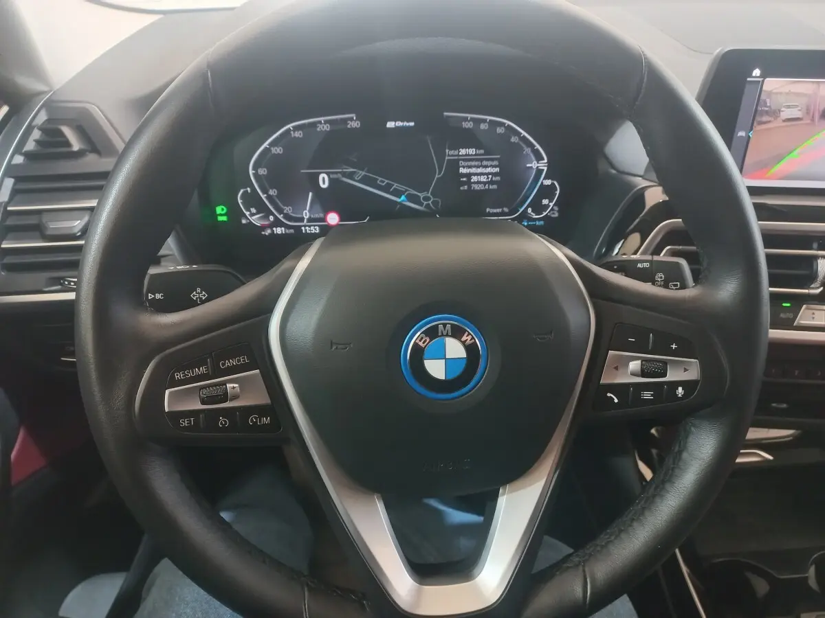 Vue intérieure centrée sur le volant cuir noir du BMW X3 xDrive30e avec tableau de bord digital et écran tactile.