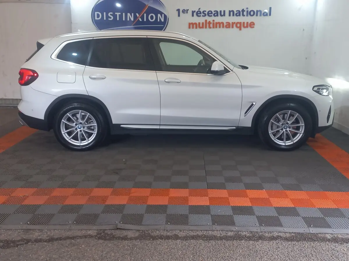 Vue de profil côté gauche d’un BMW X3 blanc hybride rechargeable garé en intérieur avec jantes alliage et feux arrière allumés.