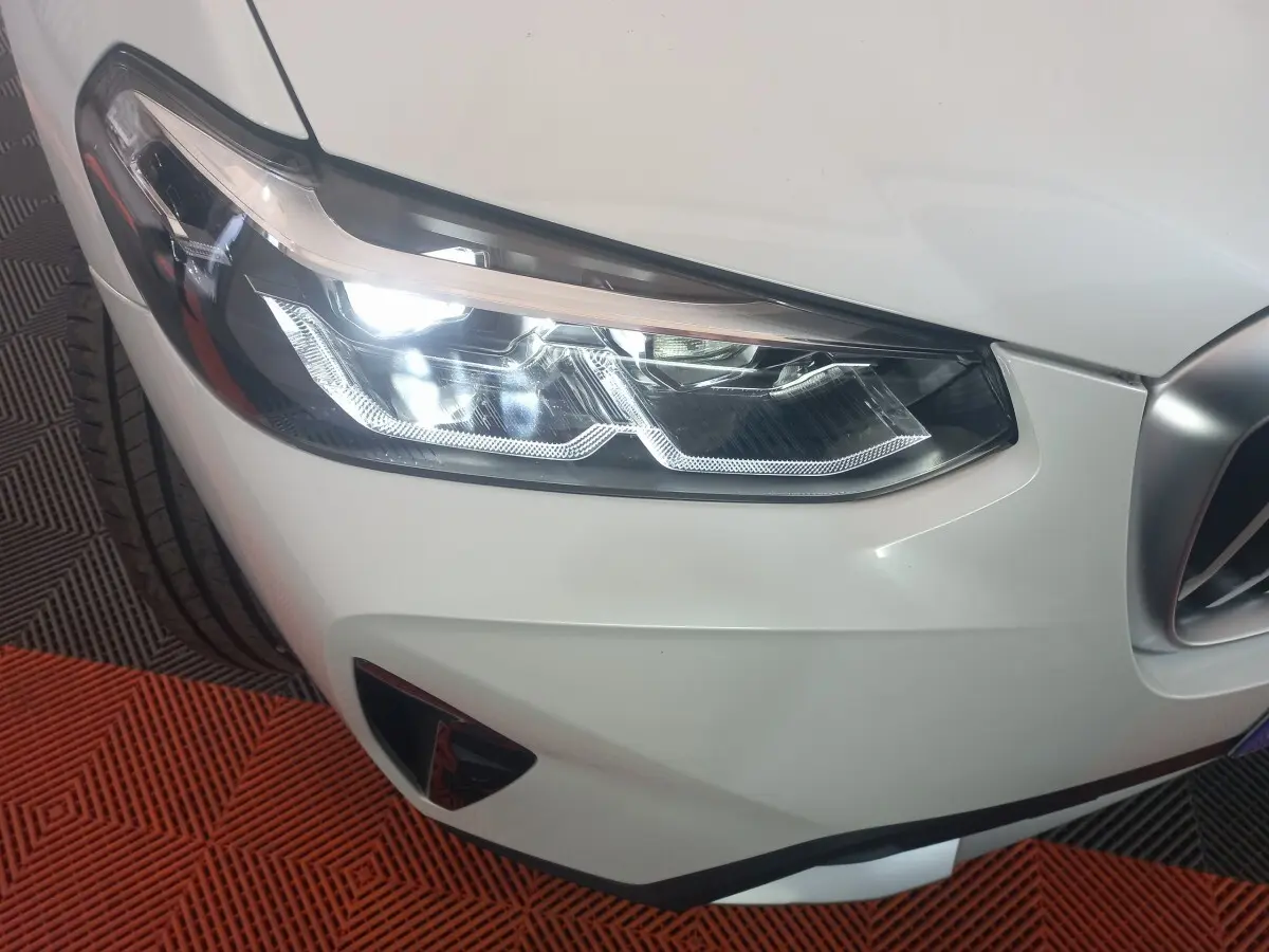 Gros plan sur le phare avant droit allumé d’un BMW X3 blanc, avec partie de la calandre visible.