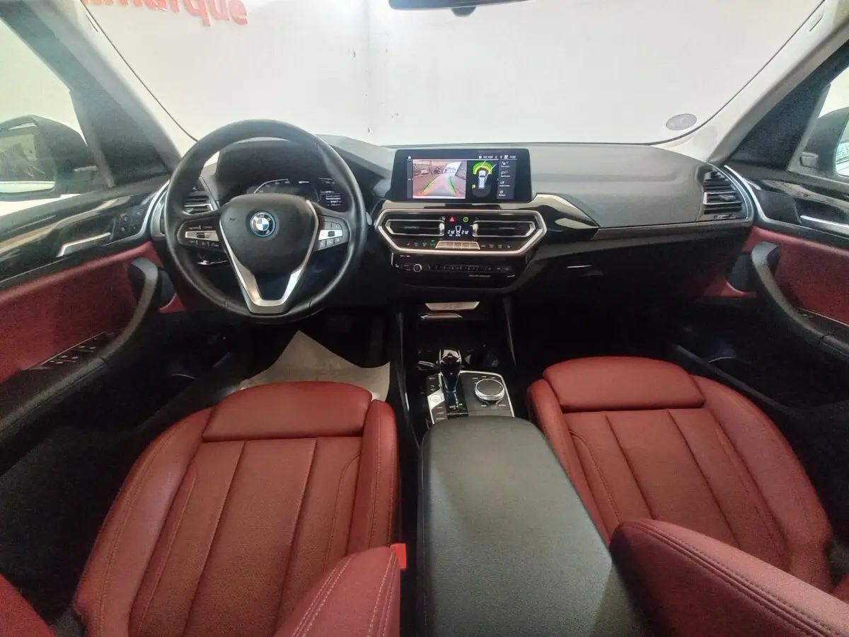 Vue intérieure avant du BMW X3 xDrive30e 2022 avec sièges cuir rouge et tableau de bord noir équipé d’un écran tactile central.