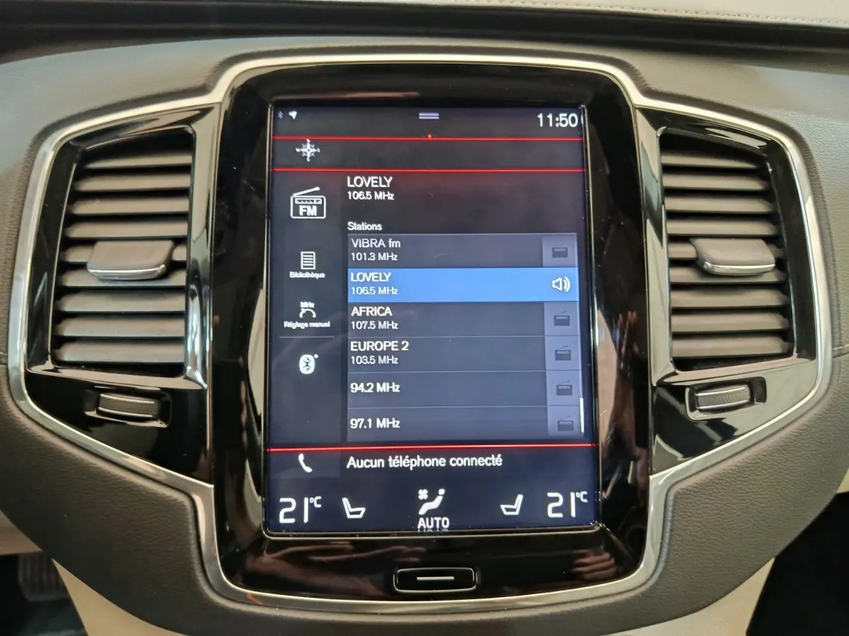 Écran tactile central du tableau de bord du Volvo XC90 gris clair, affichant les stations radio FM.