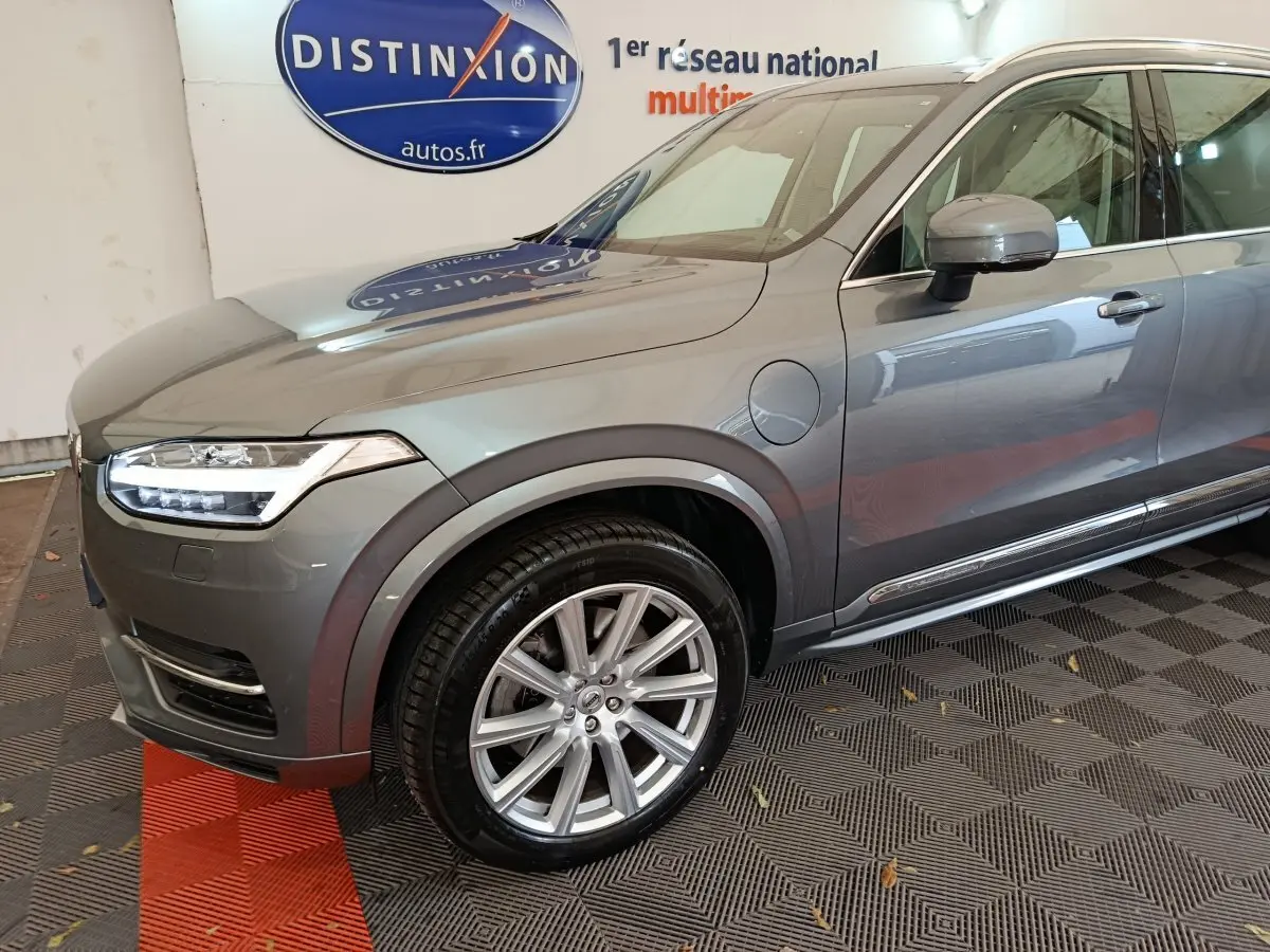 Vue 3/4 avant gauche du Volvo XC90 gris clair avec jantes alliage et prise de recharge visible sur l'aile avant.