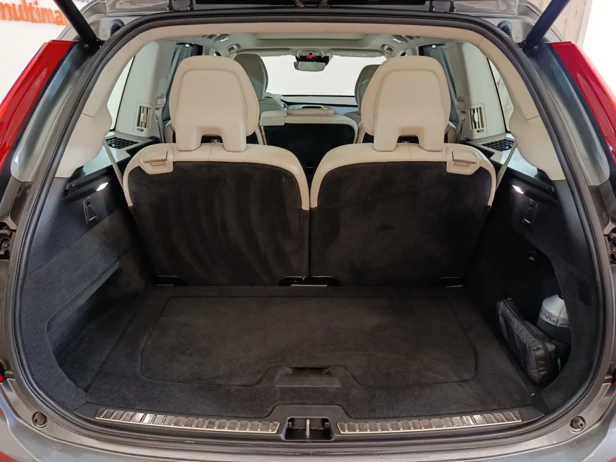 Coffre ouvert d'un Volvo XC90 gris clair, vue arrière montrant la 3ème rangée de sièges individuels beige et plancher plat.