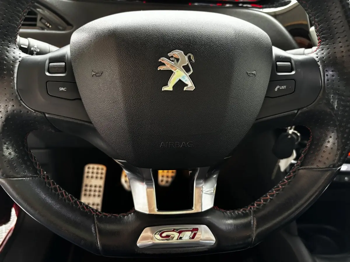 Gros plan sur le volant cuir noir perforé avec logo Peugeot et sigle GTI rouge, pédales sport en arrière-plan.