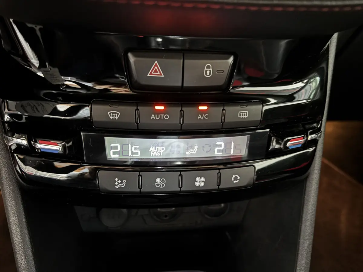 Vue rapprochée de la console centrale noire de la Peugeot 208 rouge, affichant la climatisation digitale bi-zone à 21,5°C.