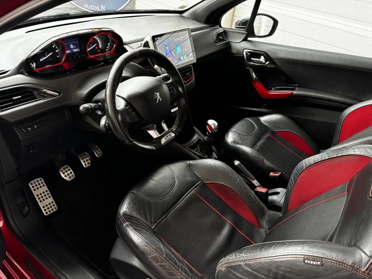 Intérieur avant de la Peugeot 208 GTI rouge, sièges cuir noir et rouge, volant GTI, tableau de bord digital et levier manuel.