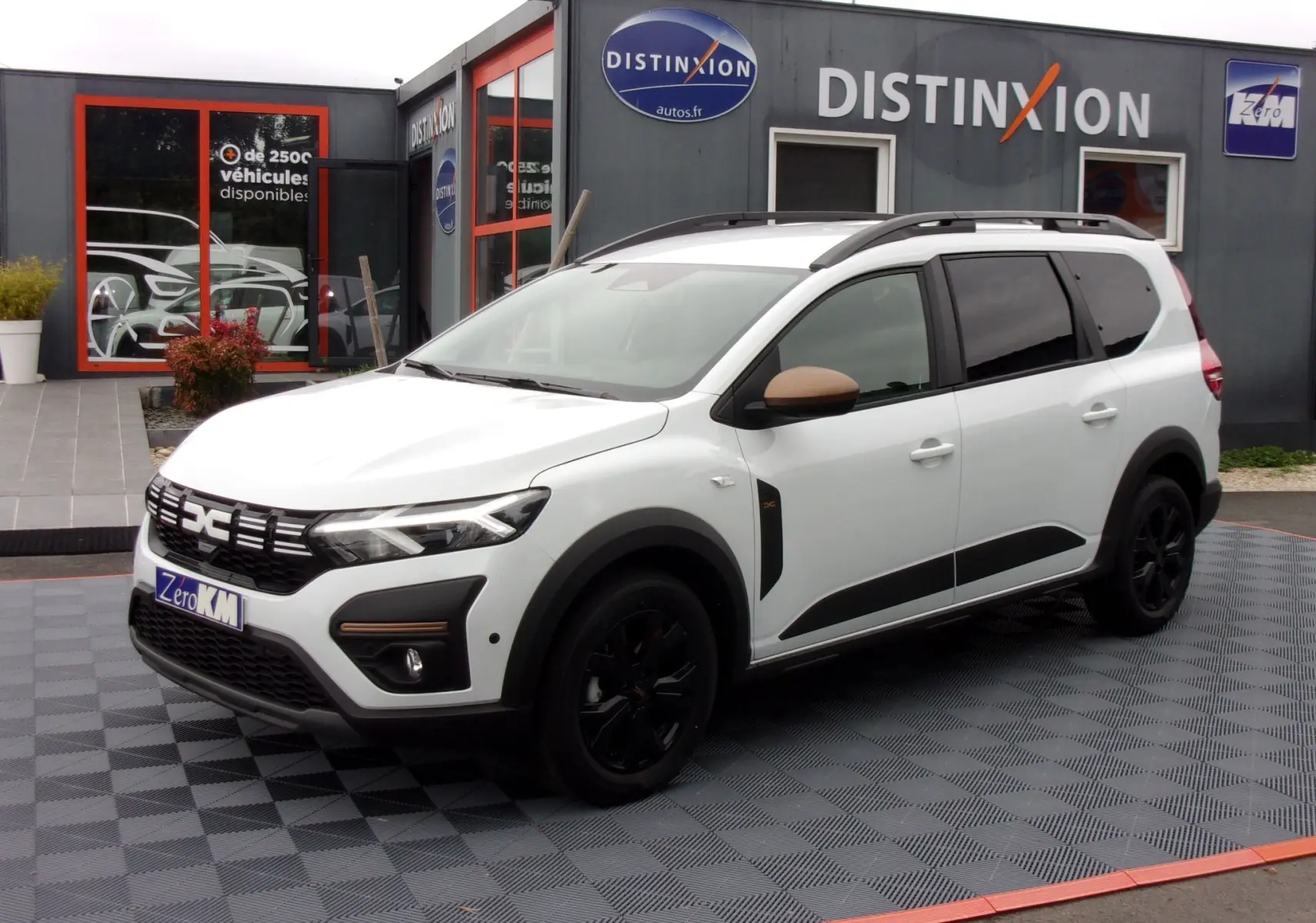 Vue 3/4 avant droit du Dacia Jogger blanc avec détails noirs et rétroviseurs marron, stationné devant un bâtiment commercial.