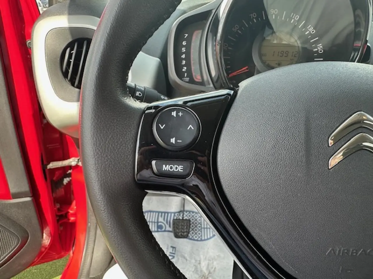 Gros plan sur le volant noir de la Citroën C1 rouge Scarlet, avec commandes audio intégrées et tableau de bord visible.