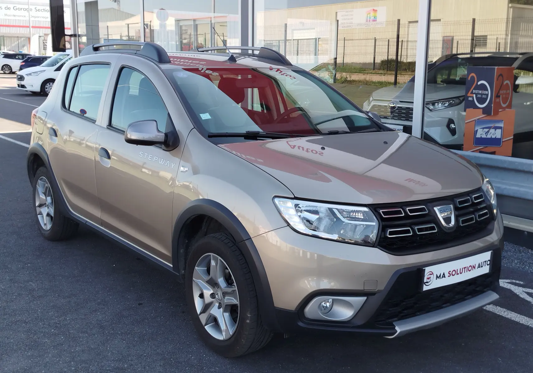 Vue 3/4 avant d'une Dacia Sandero Stepway beige dune avec barres de toit et jantes alliage, stationnée devant un garage.