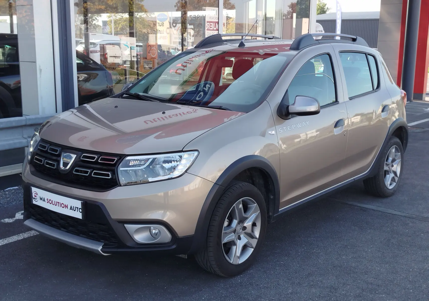 Dacia Sandero Stepway beige dune vue 3/4 avant droit avec barres de toit et jantes alliage.