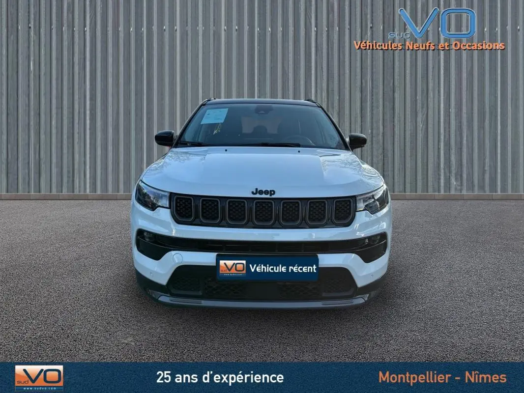 Vue frontale d'un Jeep Compass blanc 2023 avec calandre noire et phares LED, stationné devant un mur métallique.