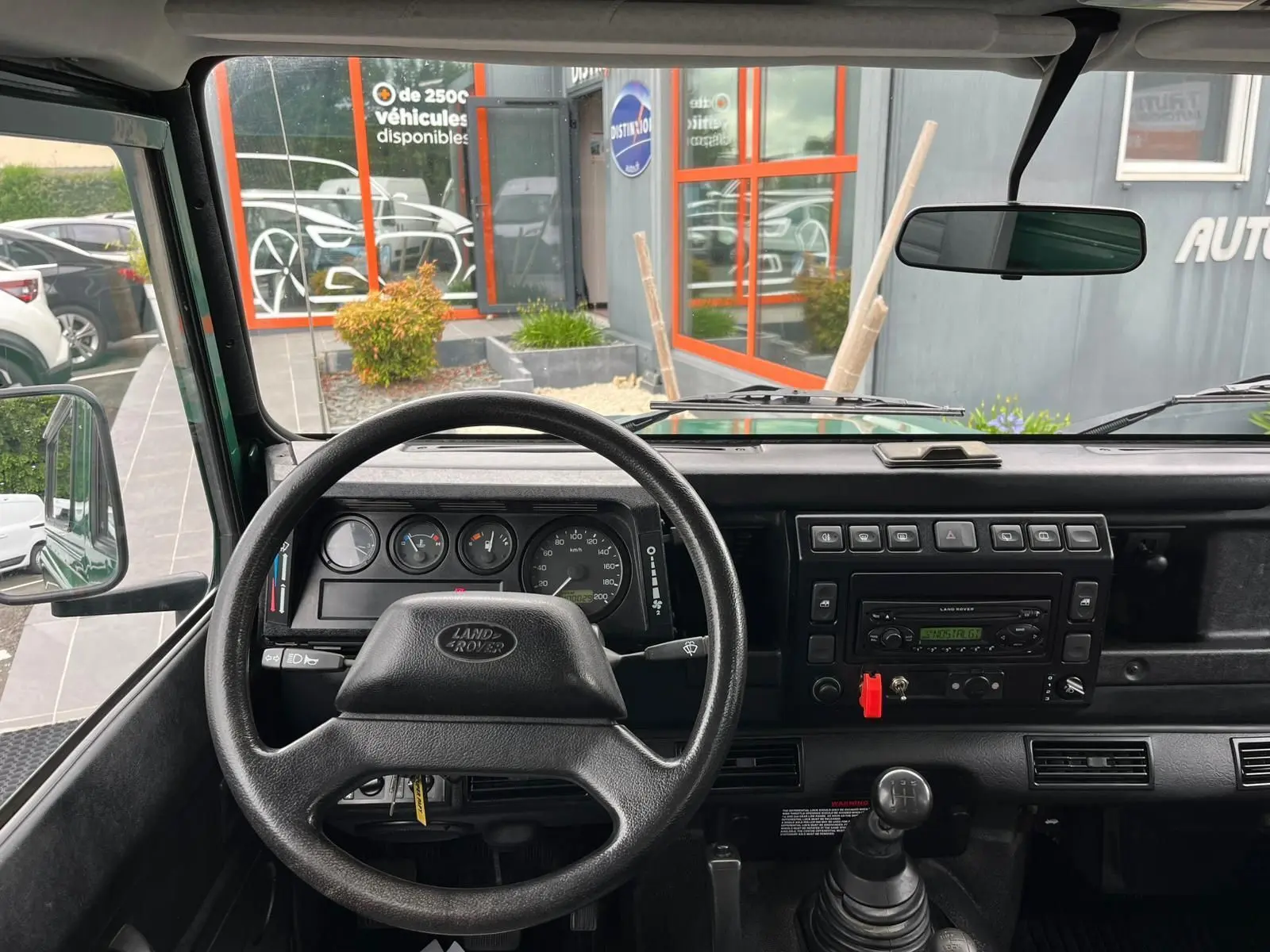 Vue intérieure frontale du tableau de bord noir du Land Rover Defender 90 vert, avec volant, compteurs et levier de vitesses manuel.