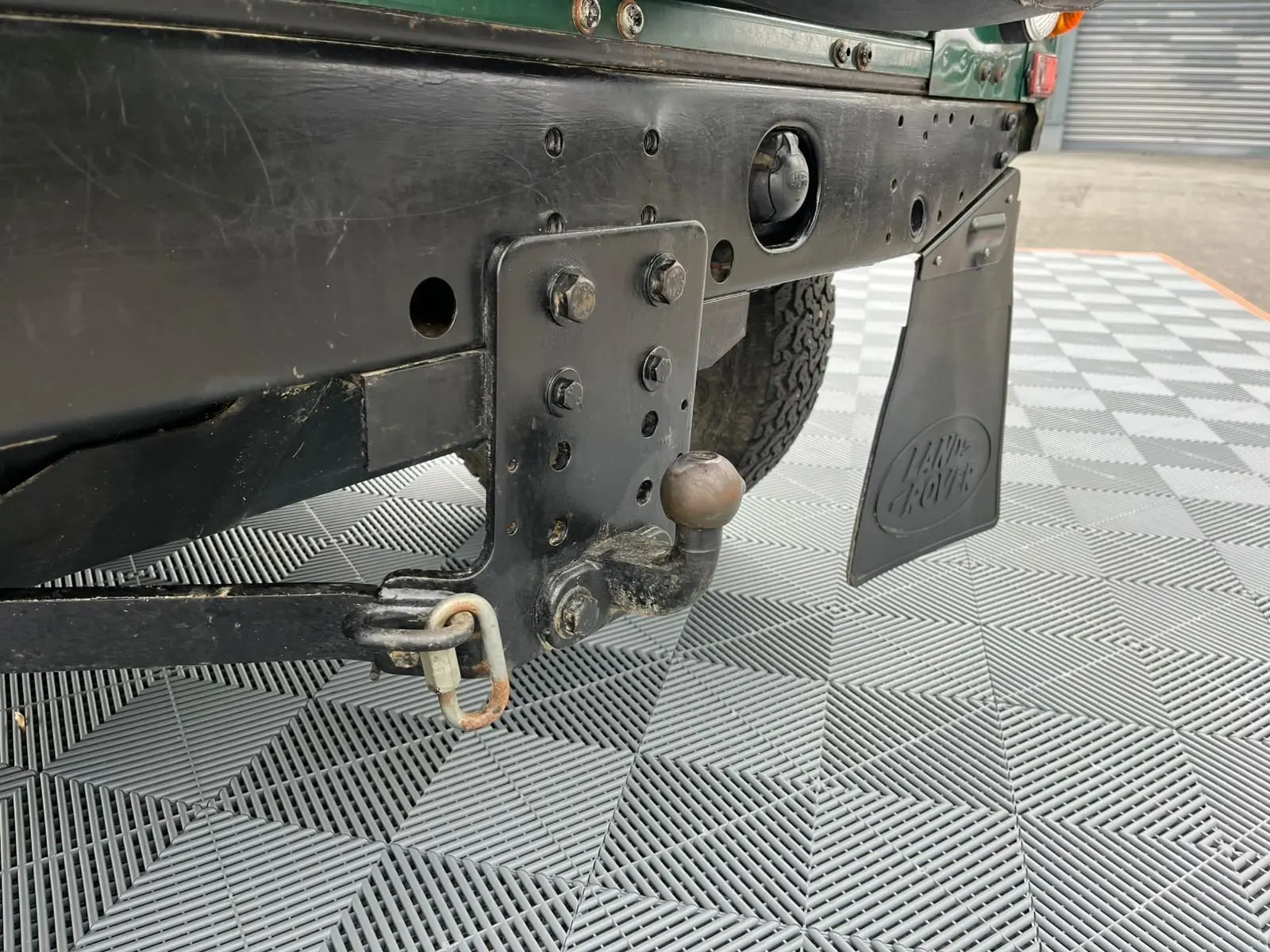 Gros plan sur l'attelage arrière noir et le bavette Land Rover d'un Defender 90 vert, vue côté gauche.