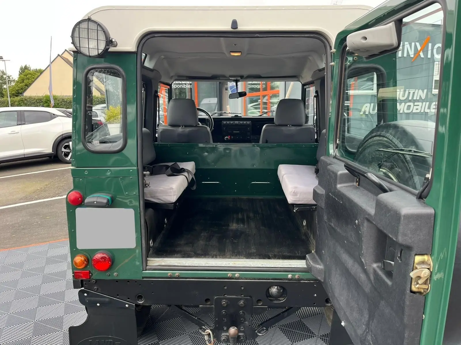 Vue arrière ouverte du Land Rover Defender 90 vert 2004 montrant l'intérieur utilitaire avec banquettes latérales et roue de secours.