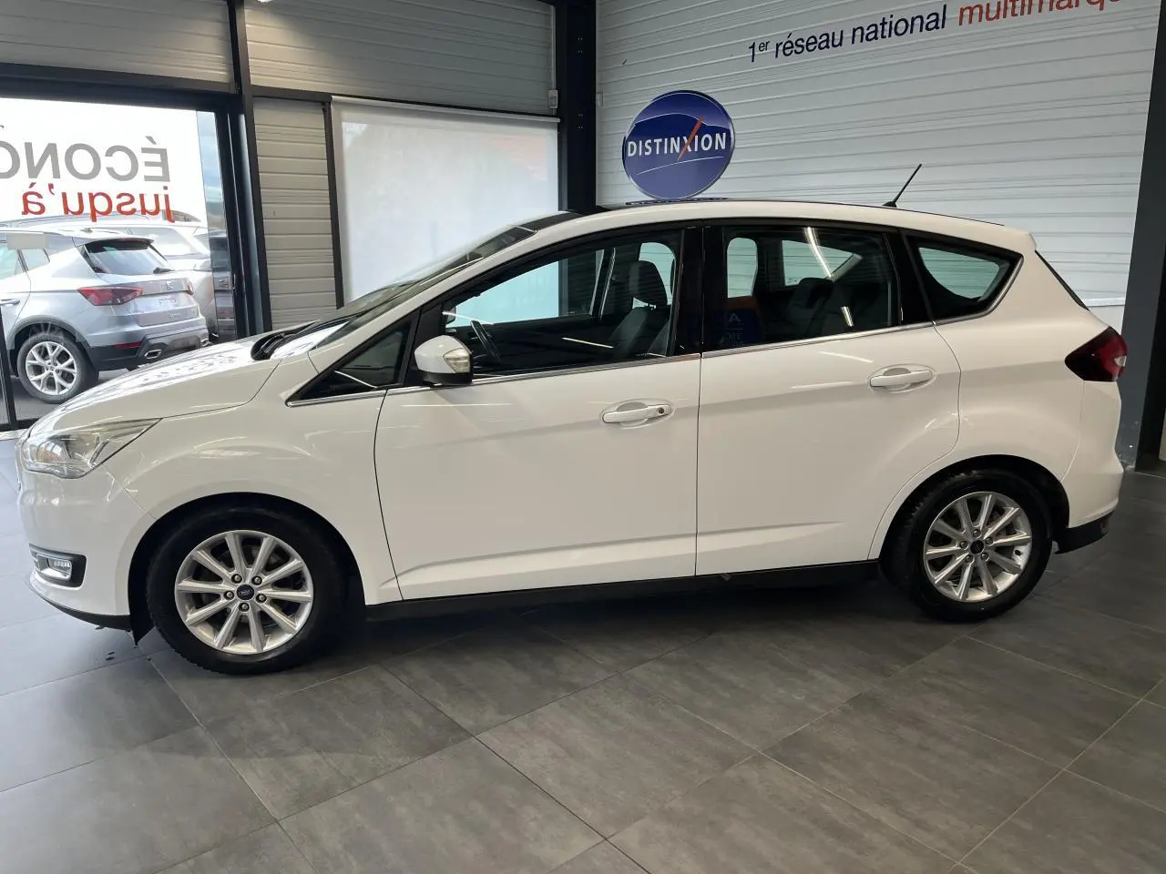Ford C-MAX blanc vu de profil côté gauche dans un showroom avec jantes alliage et toit panoramique.