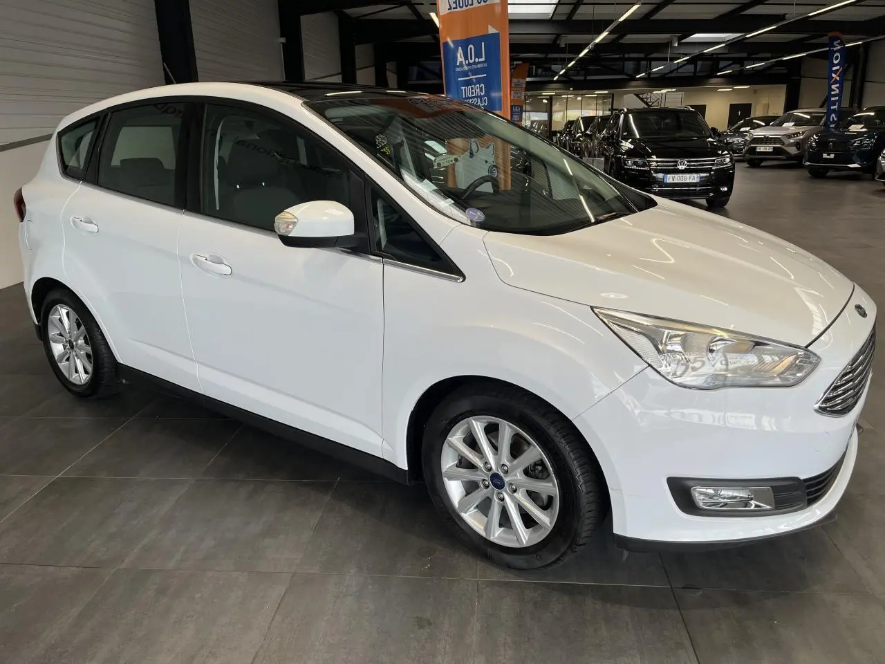 Vue 3/4 avant droit d'une Ford C-MAX blanche avec jantes alliage et toit panoramique en showroom.