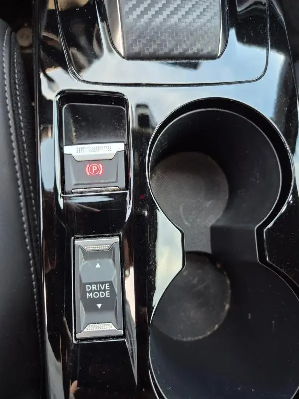 Gros plan sur la console centrale noire brillante du Peugeot 2008 Hybrid 145 e-DCS6 GT avec bouton frein à main et sélection drive mode.
