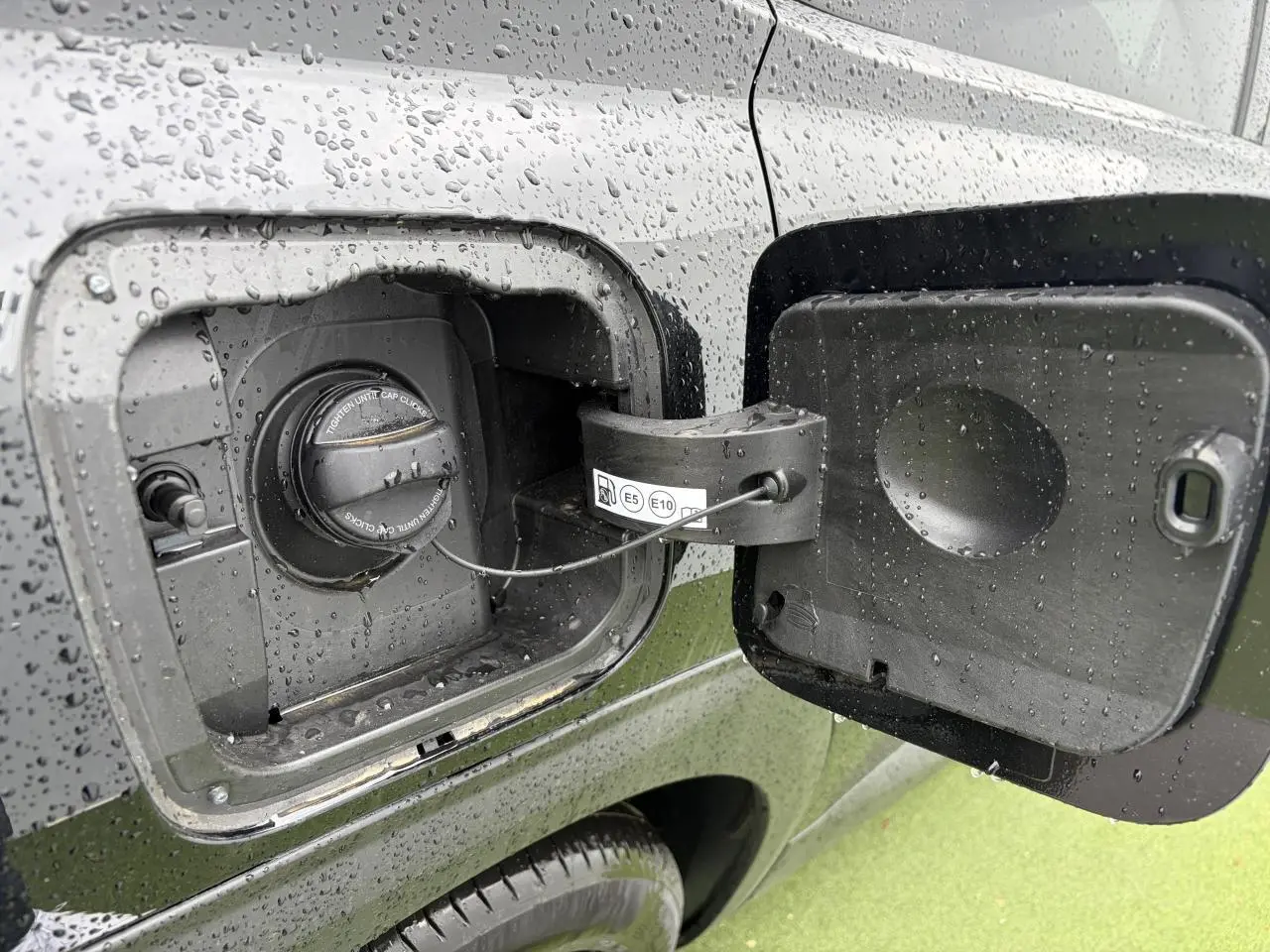 Gros plan sur la trappe de carburant ouverte du Citroën C5 Aircross noir métal, avec bouchon essence sous la pluie.