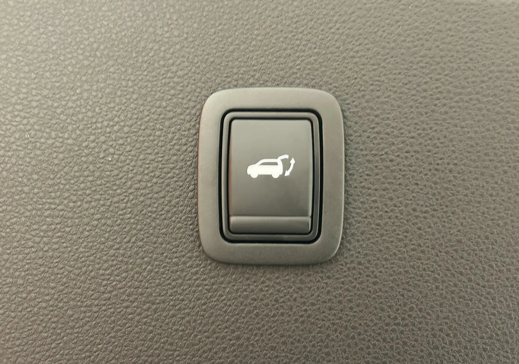 Bouton intérieur noir avec symbole blanc d'ouverture électrique du hayon sur fond de texture plastique grise.
