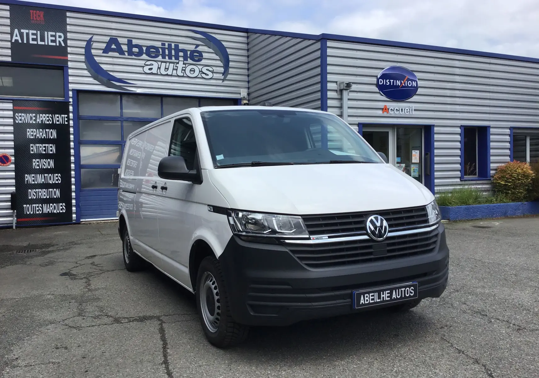 Vue 3/4 avant d'un Volkswagen Transporter 6.1 blanc stationné devant un garage avec logo Abeilhé Autos.