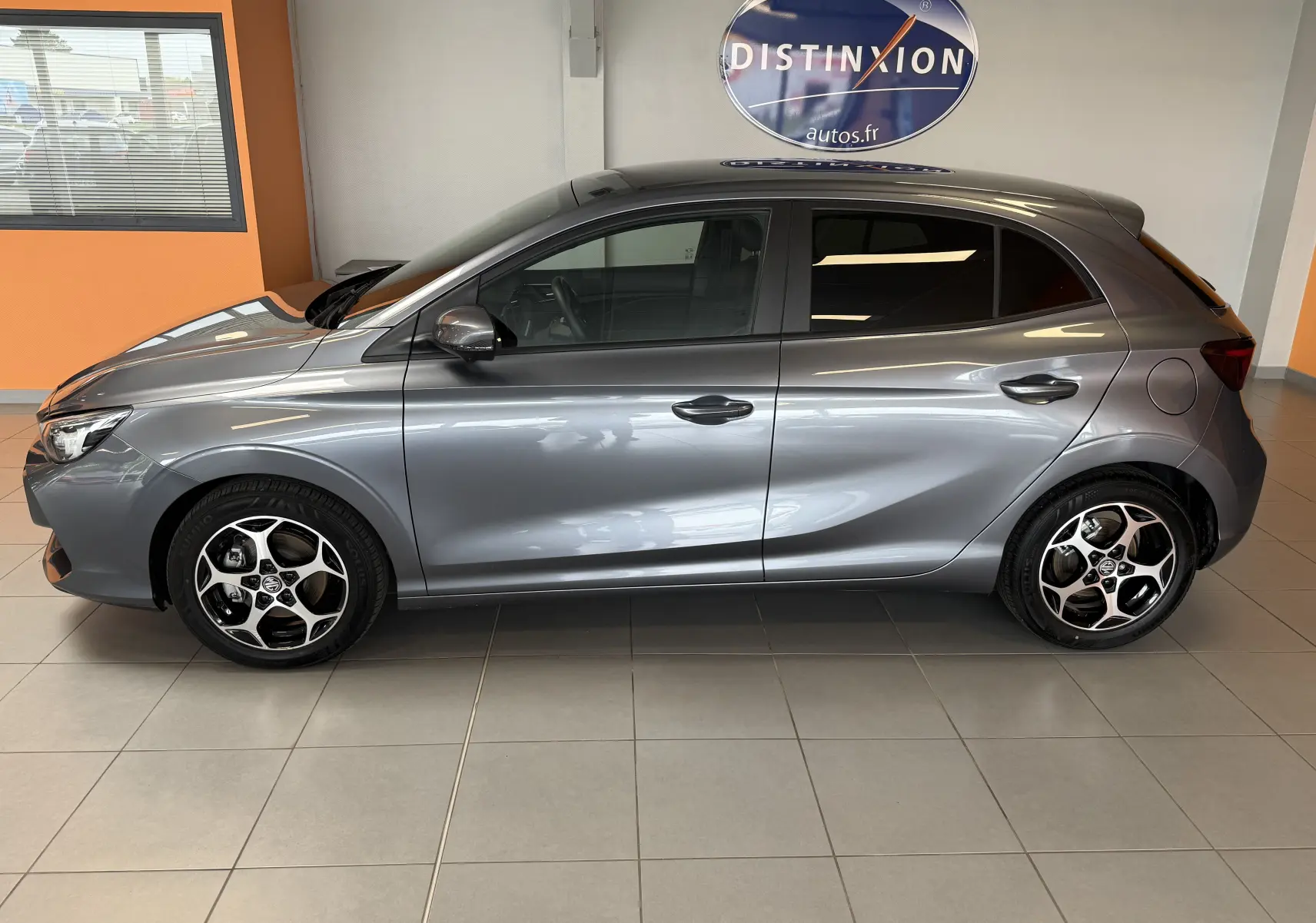 Vue de profil côté gauche d'une MG3 hybride gris métallisé avec jantes bi-ton et vitres surteintées en intérieur showroom.