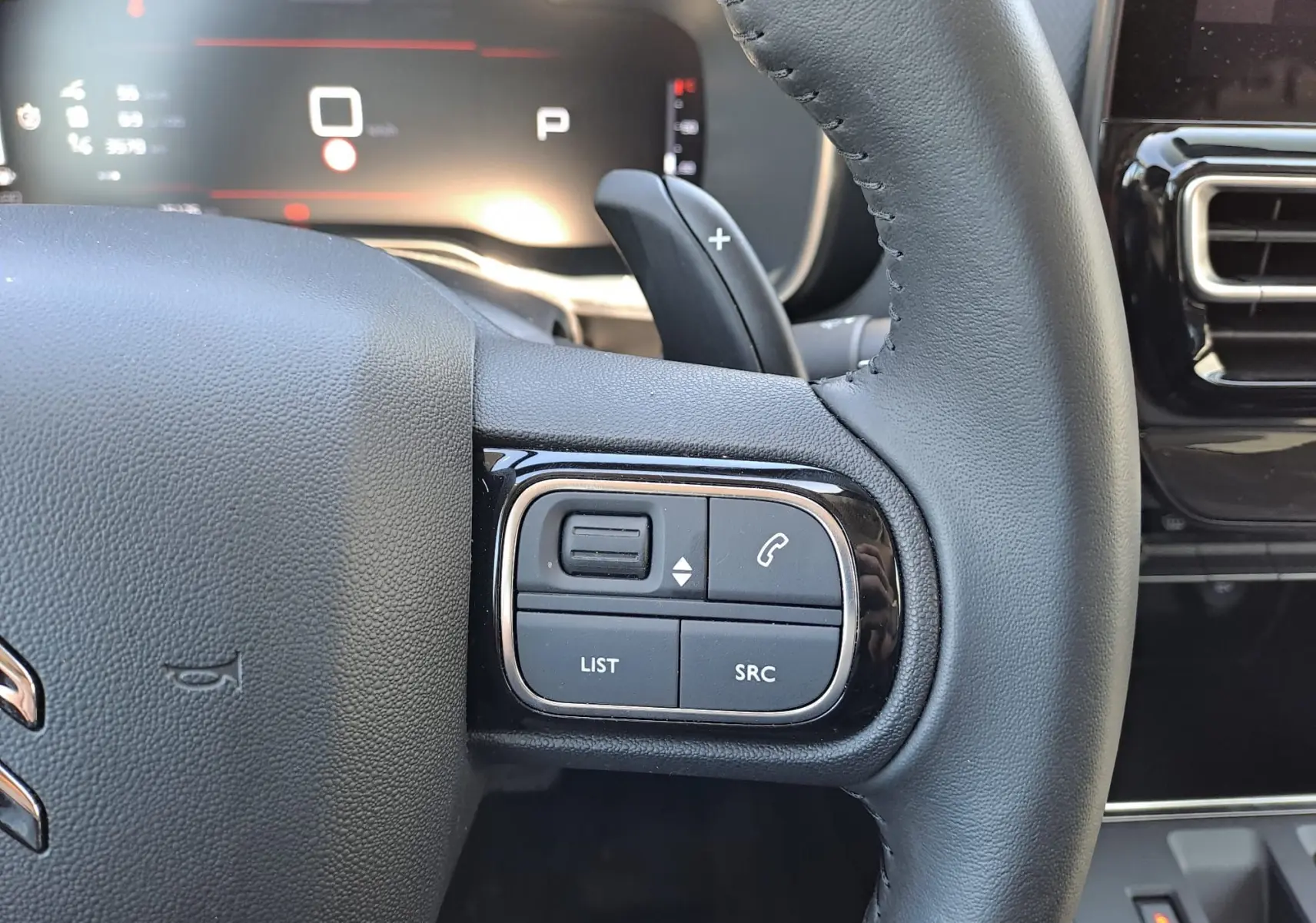 Gros plan sur les commandes gauche du volant cuir du Citroën C5 Aircross 2024 hybride, avec tableau de bord numérique en arrière-plan.