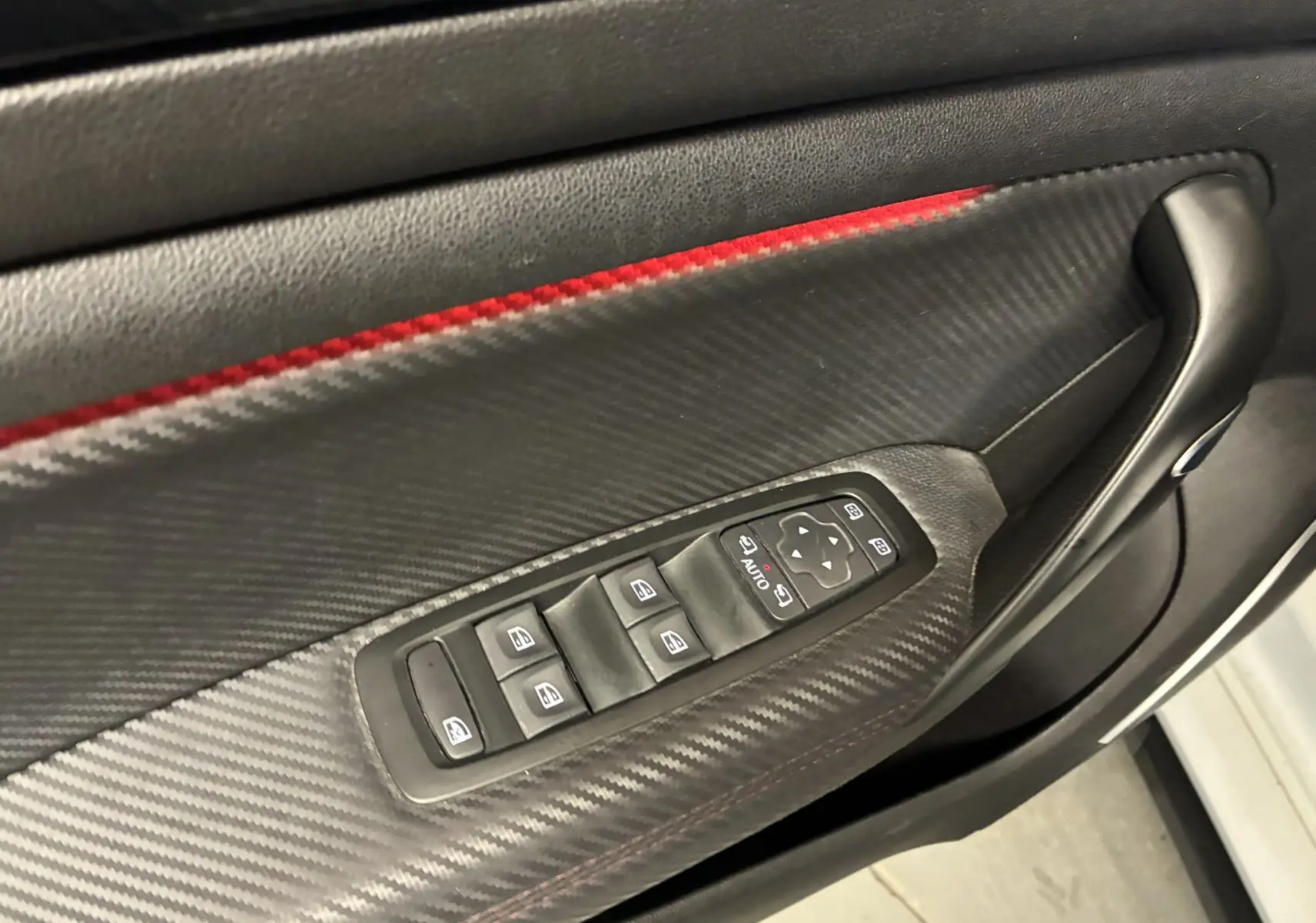 Gros plan sur la commande des vitres électriques côté gauche avec finition carbone et surpiqûres rouges dans l'habitacle noir.