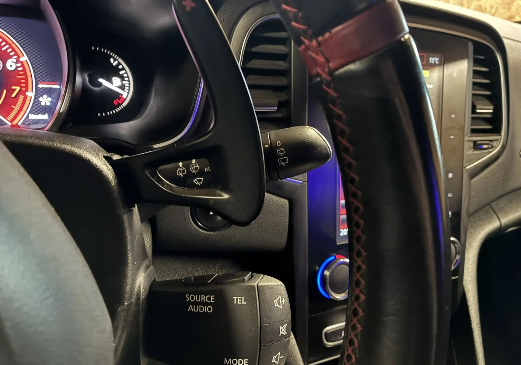 Gros plan sur le volant noir avec surpiqûres rouges et commandes audio, tableau de bord et écran central en arrière-plan.